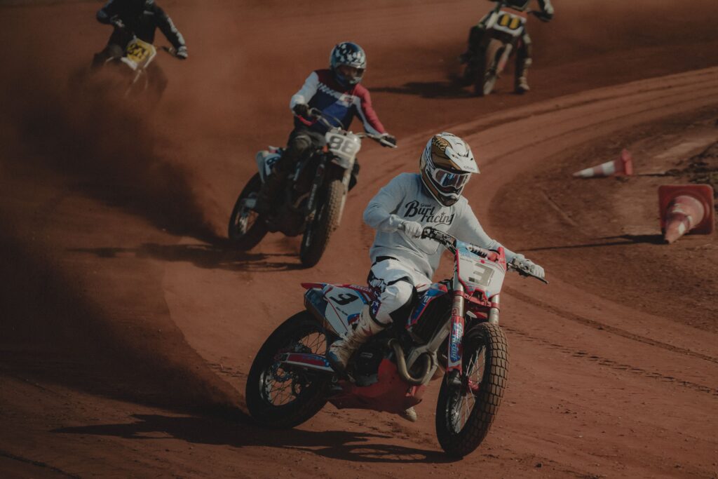 Czterech zawodników flat track pokonuje zakręt na motocyklach podczas wyścigu na ziemnym torze.